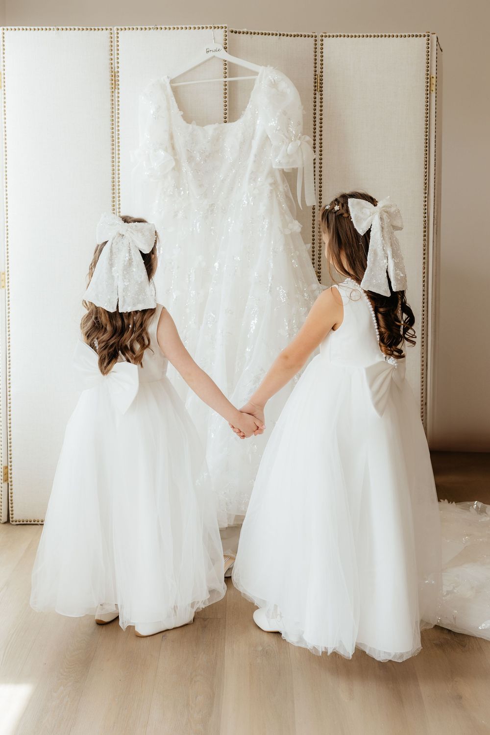 Two flower girls in white dresses holding hands while admiring a wedding gown hanging on a screen.