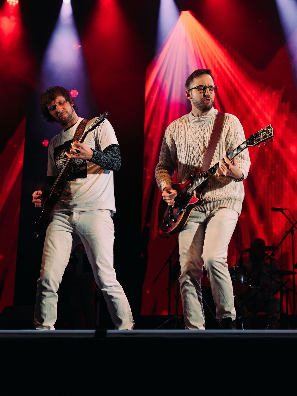 Deux guitaristes vêtus de blanc jouent sur scène sous un éclairage théâtral rouge. Florent Vause