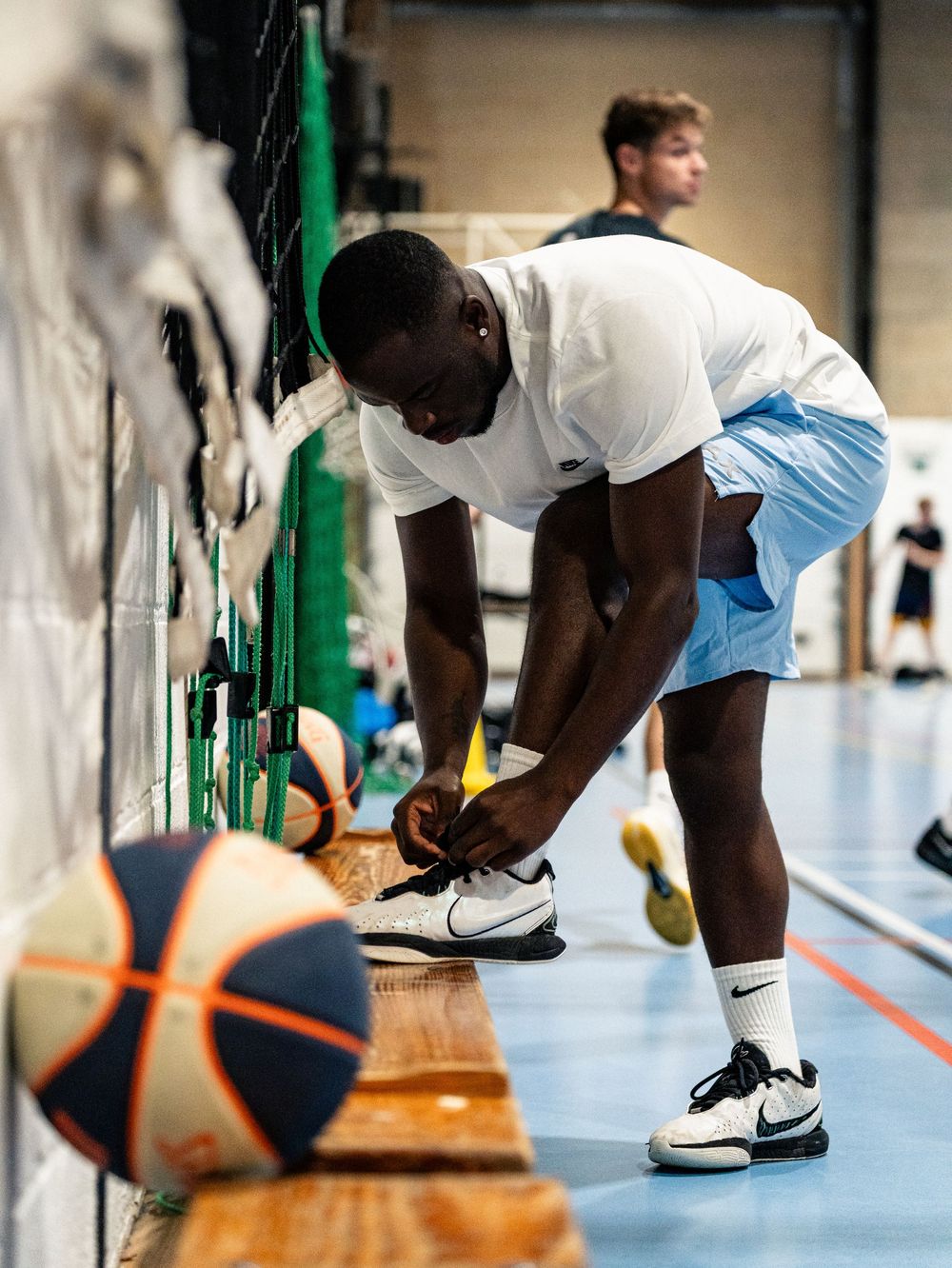 Joueur de basket-ball nouant ses lacets sur un terrain couvert, avec un ballon au premier plan. Florent Vause