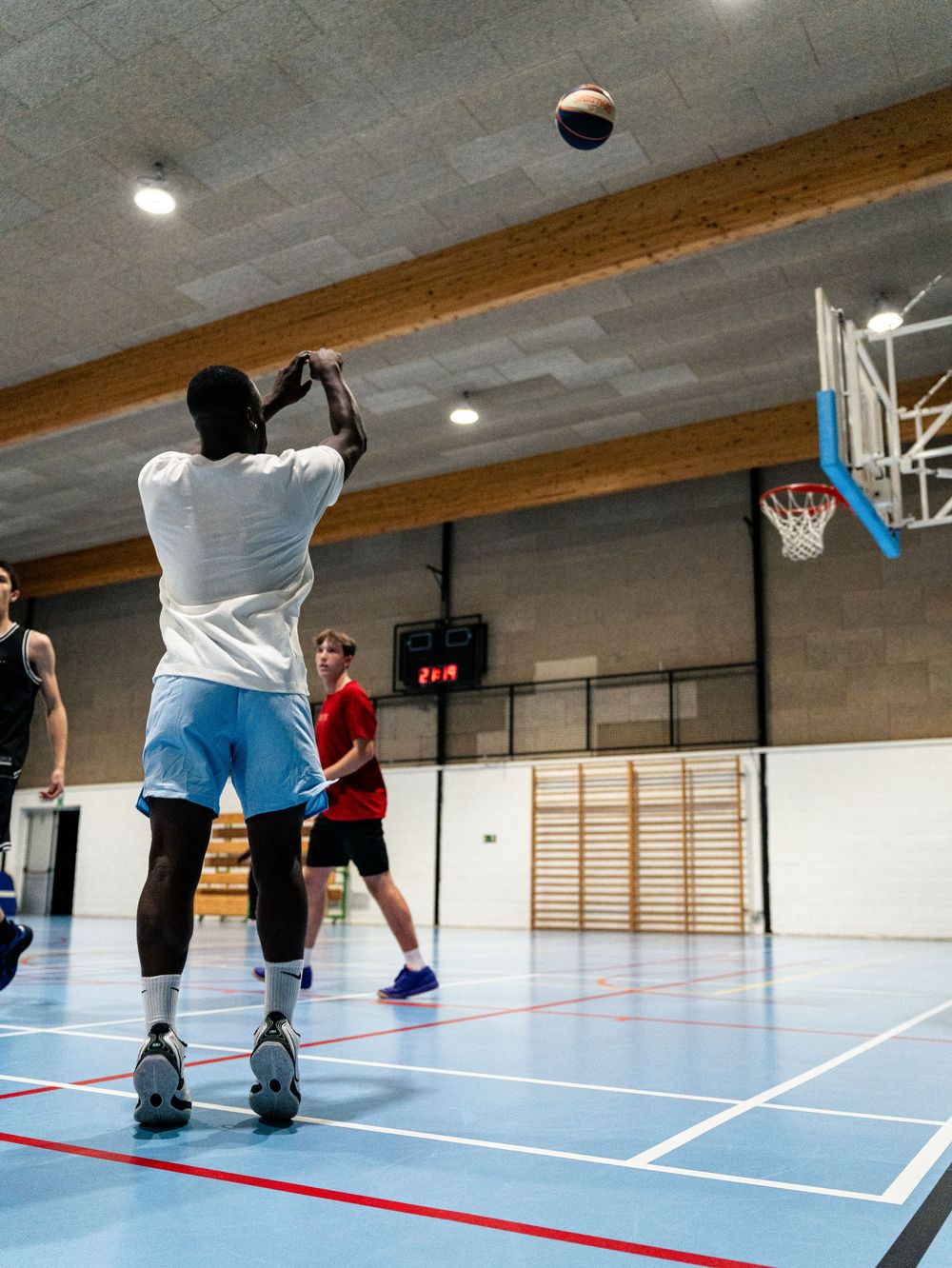 Joueur effectuant un tir en suspension sur un terrain de basket intérieur avec un panier en arrière-plan. Florent Vause