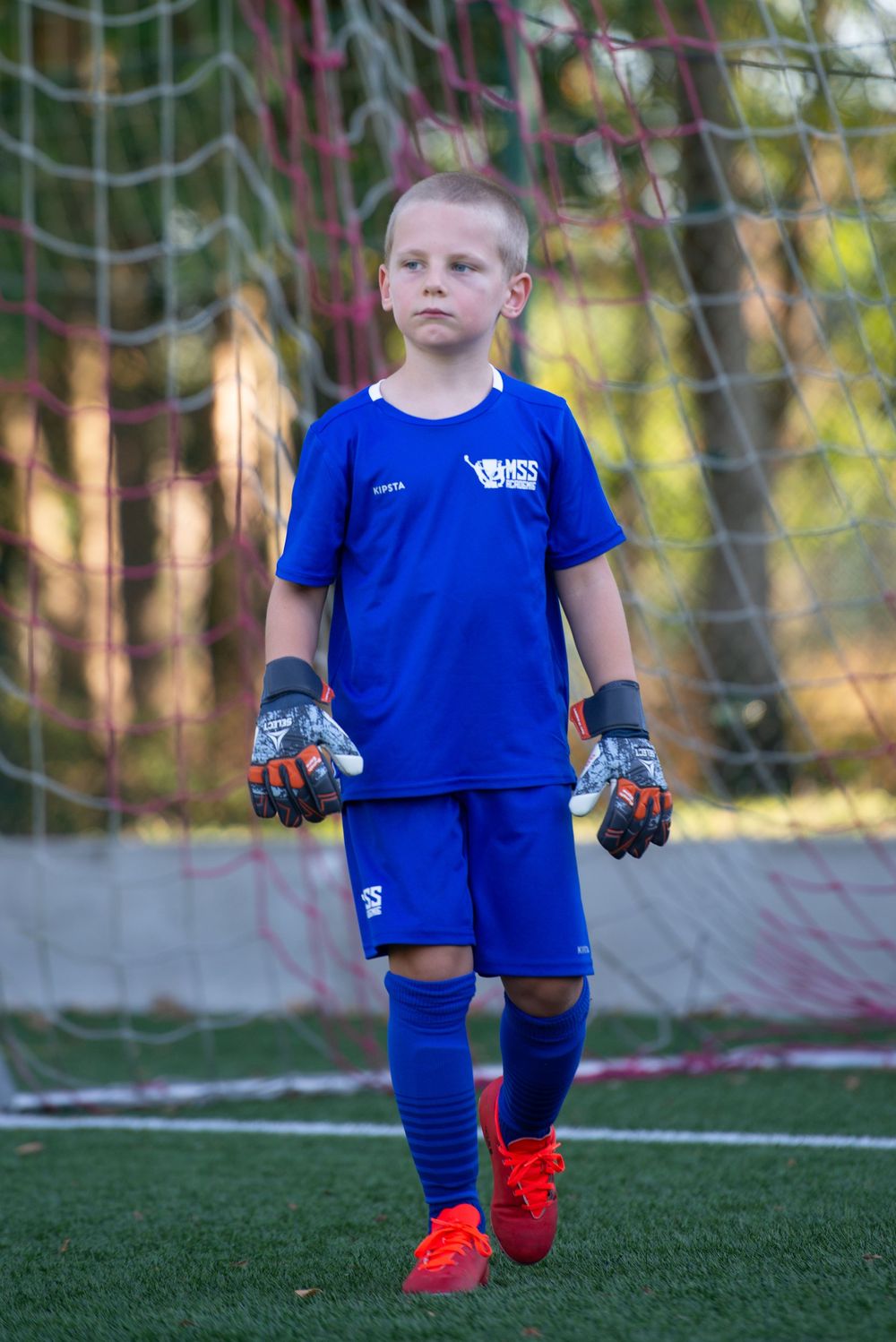 Jeune gardien de but de football vêtu d'un maillot bleu et de crampons orange se tient avec assurance devant les cages. Florent Vause