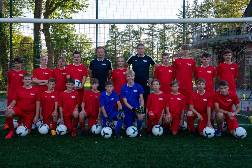 Une équipe de football junior vêtue d'uniformes rouges et bleus pose avec ses entraîneurs sur un terrain en gazon synthétique. Florent Vause
