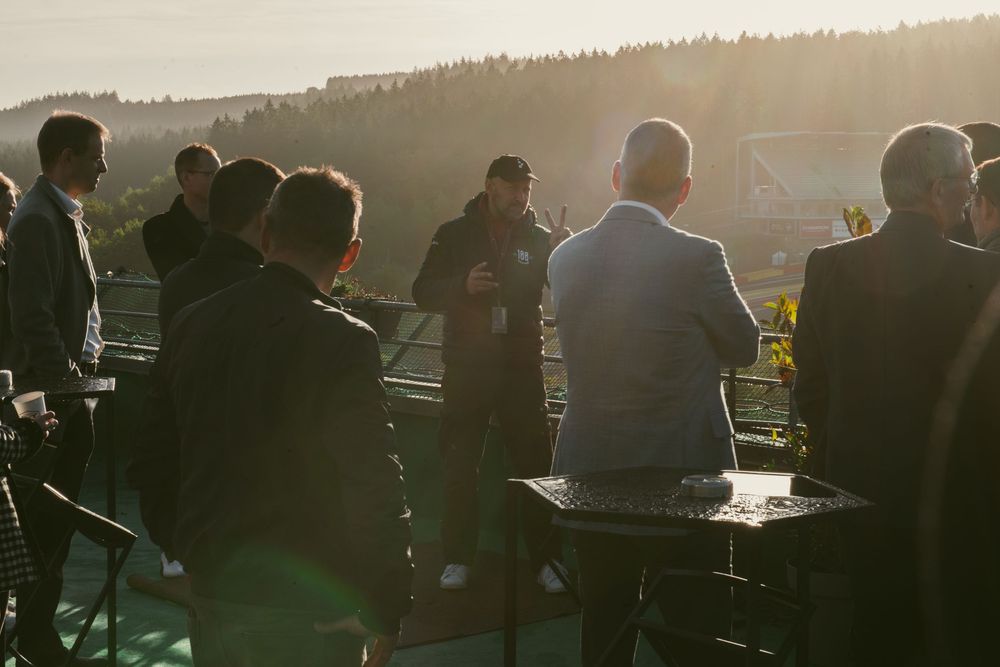 Groupe de personnes silhouettées rassemblées autour de tables à l'extérieur au coucher du soleil. Florent Vause