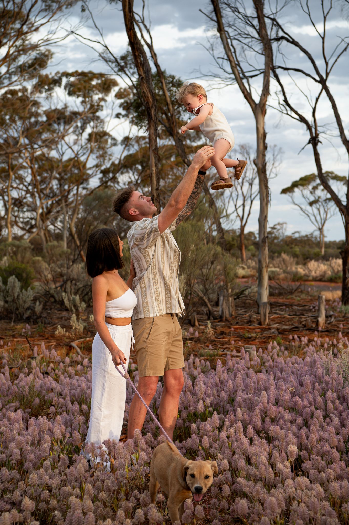 Laughing small child in white romper laying on brown blanket surrounded by purple wildflowers in outdoor setting.