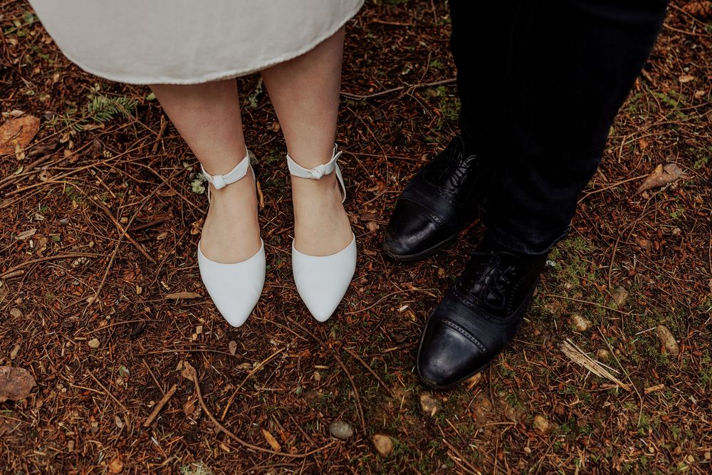 Close-up photo of formal footwear showing white pointed flats next to black dress shoes on autumn leaves.