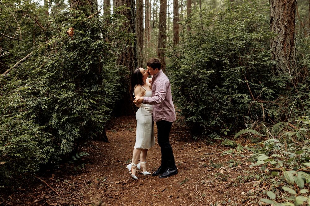 A couple shares an intimate moment during their woodland engagement photo session among tall evergreen trees.