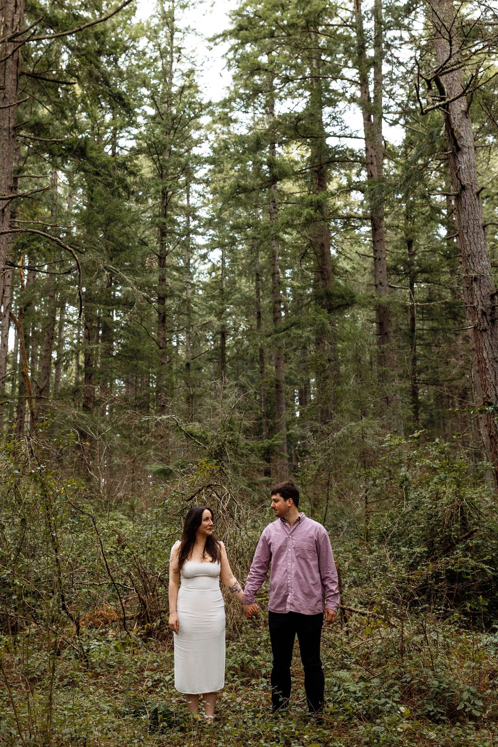 A couple walks and looks at each other lovingly in a serene forest setting with tall evergreen trees.