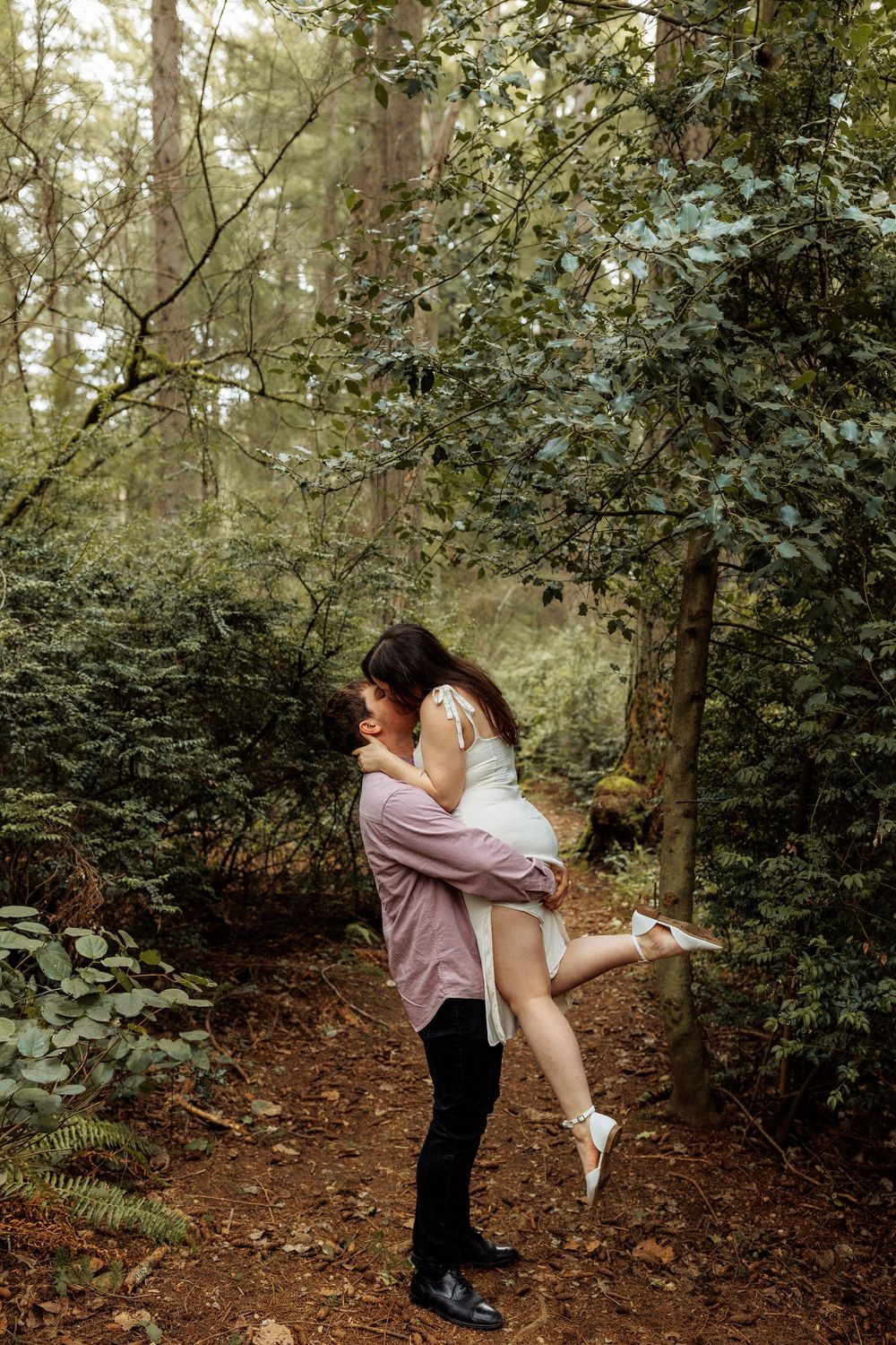 A romantic couple embraces on a forest path amidst lush greenery and filtered sunlight.