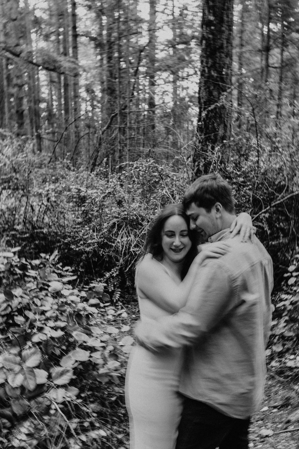 A romantic black and white photograph of a couple embracing in a moody Pacific Northwest forest setting.