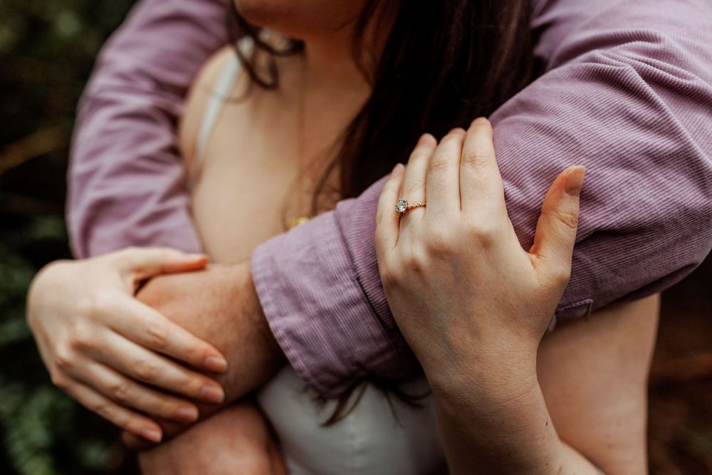 Close up of hands with engagement ring embracing in purple jacket sleeves against greenery background.