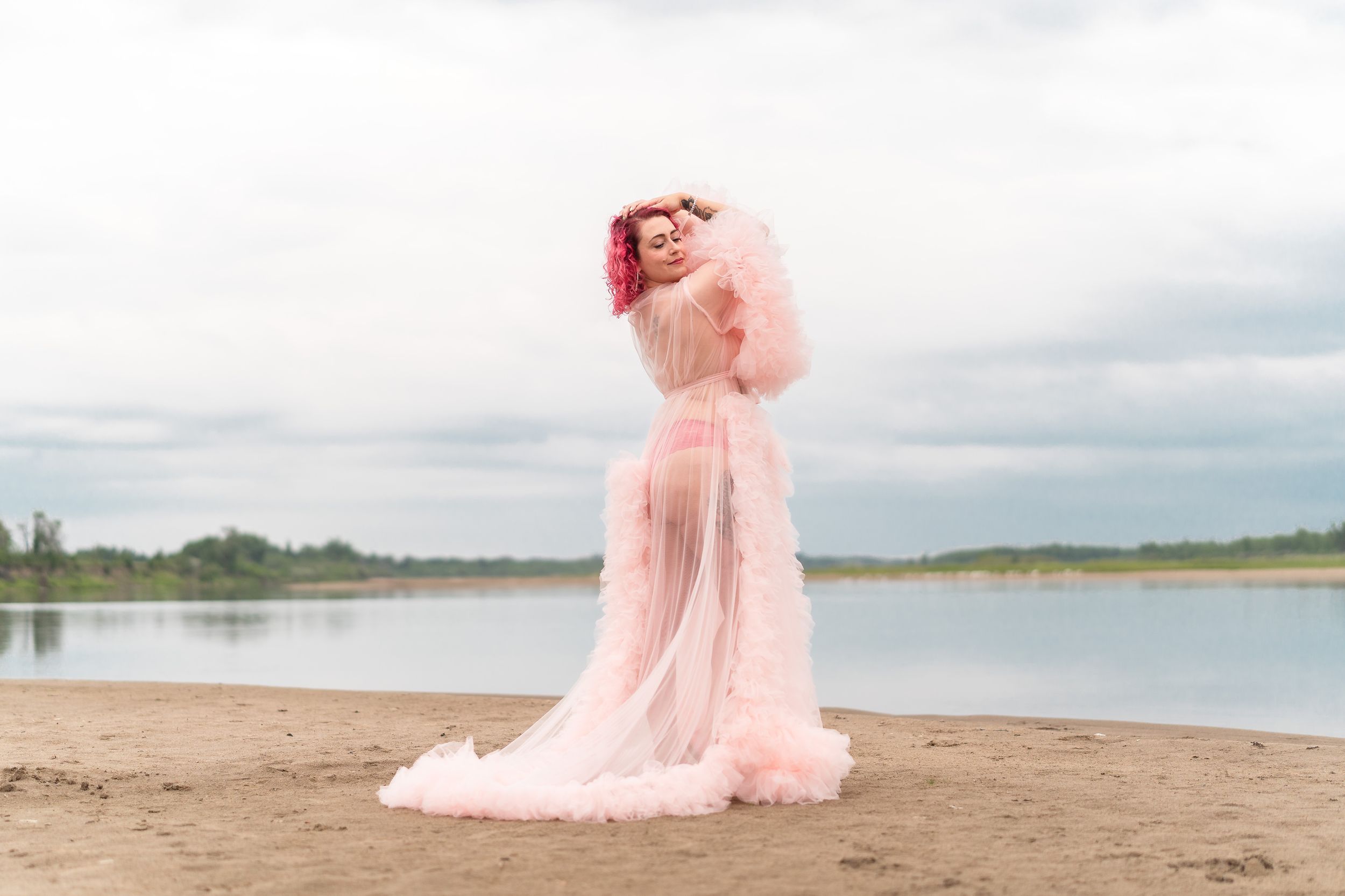 A dreamy figure in a flowing pink tulle gown poses gracefully on a beach beside a tranquil lake.