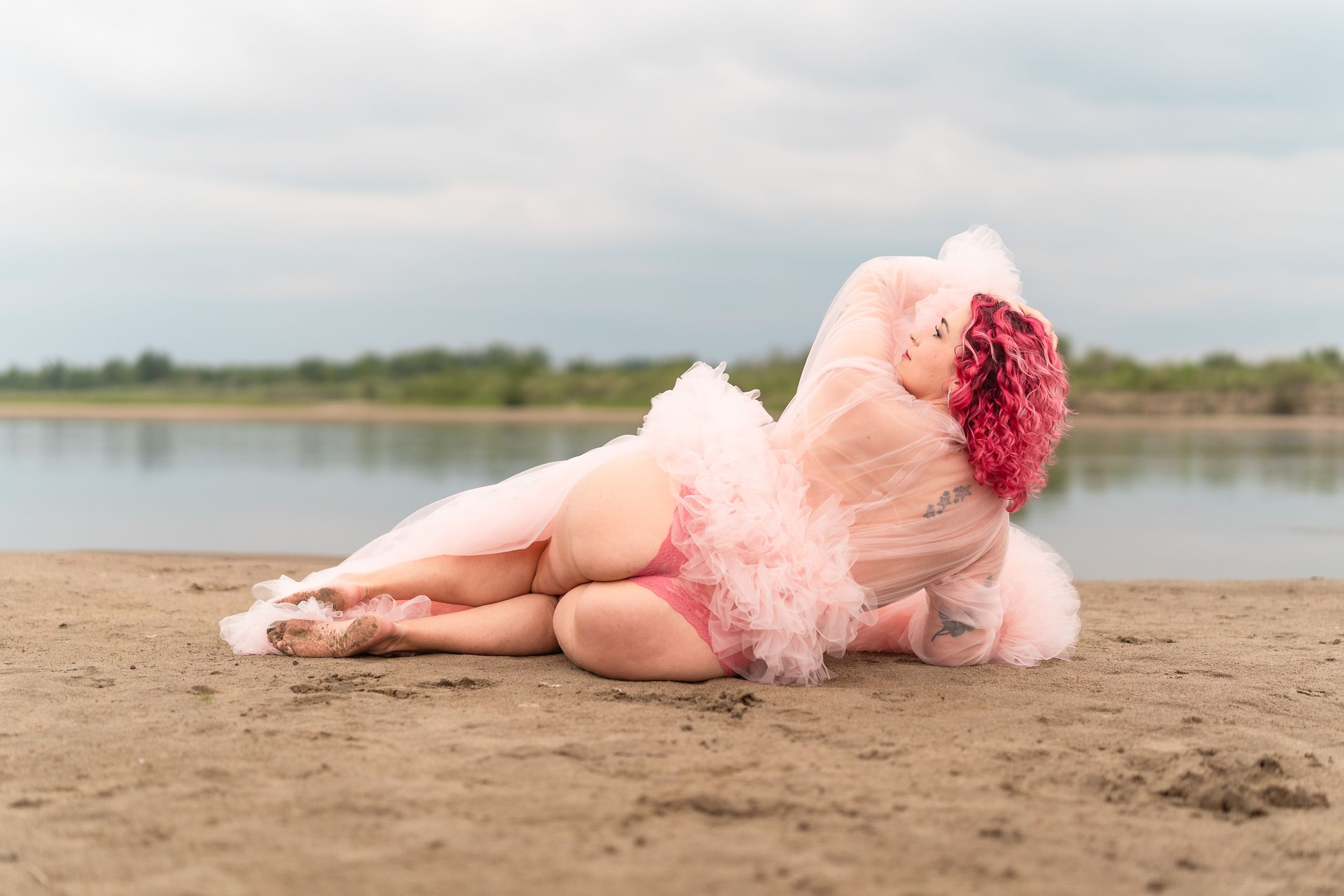 Someone in a pink tulle dress reclines on a sandy beach near calm waters at sunset.