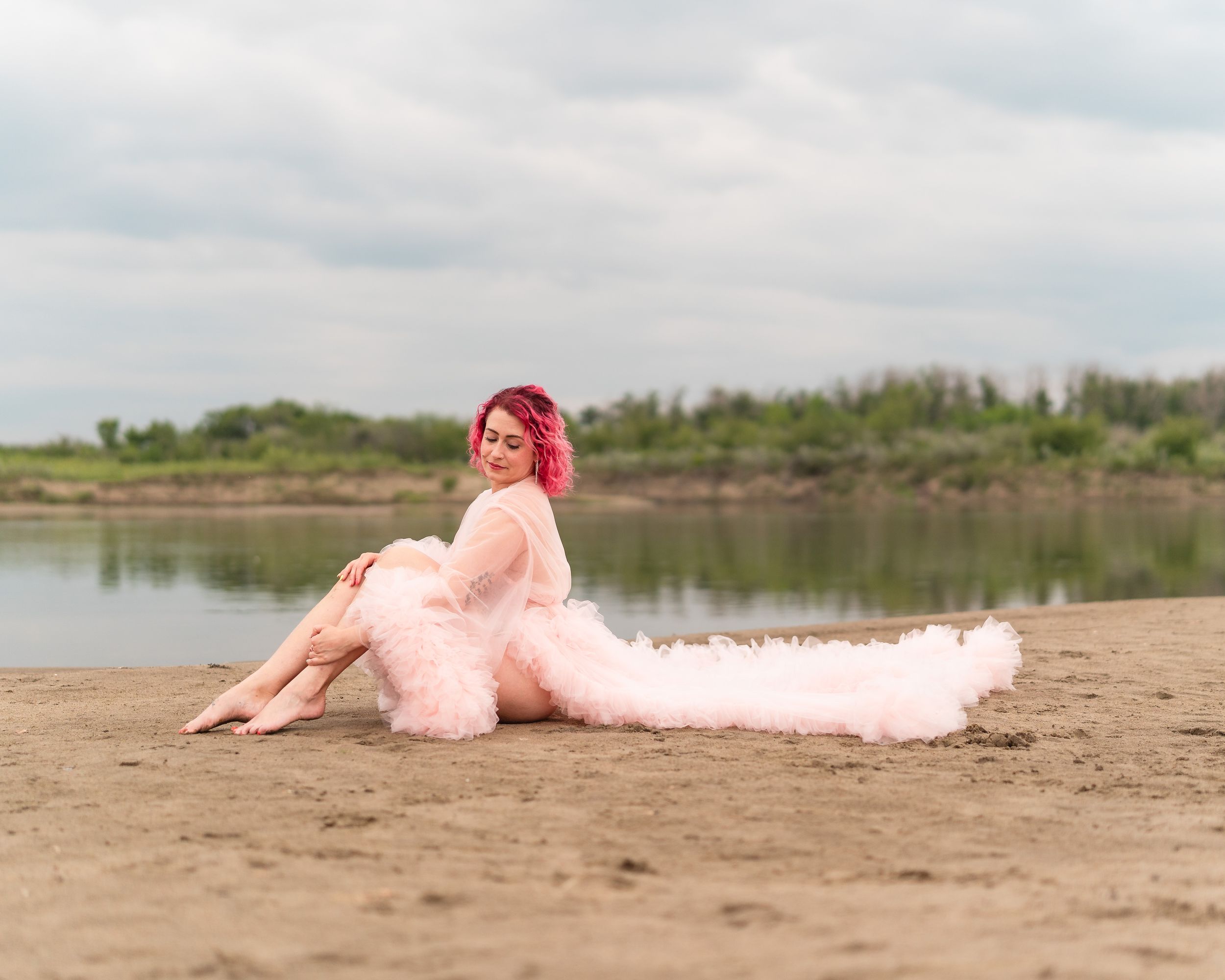 A figure in a flowing pink dress reclines on a sandy beach beside a serene lake on a cloudy day.