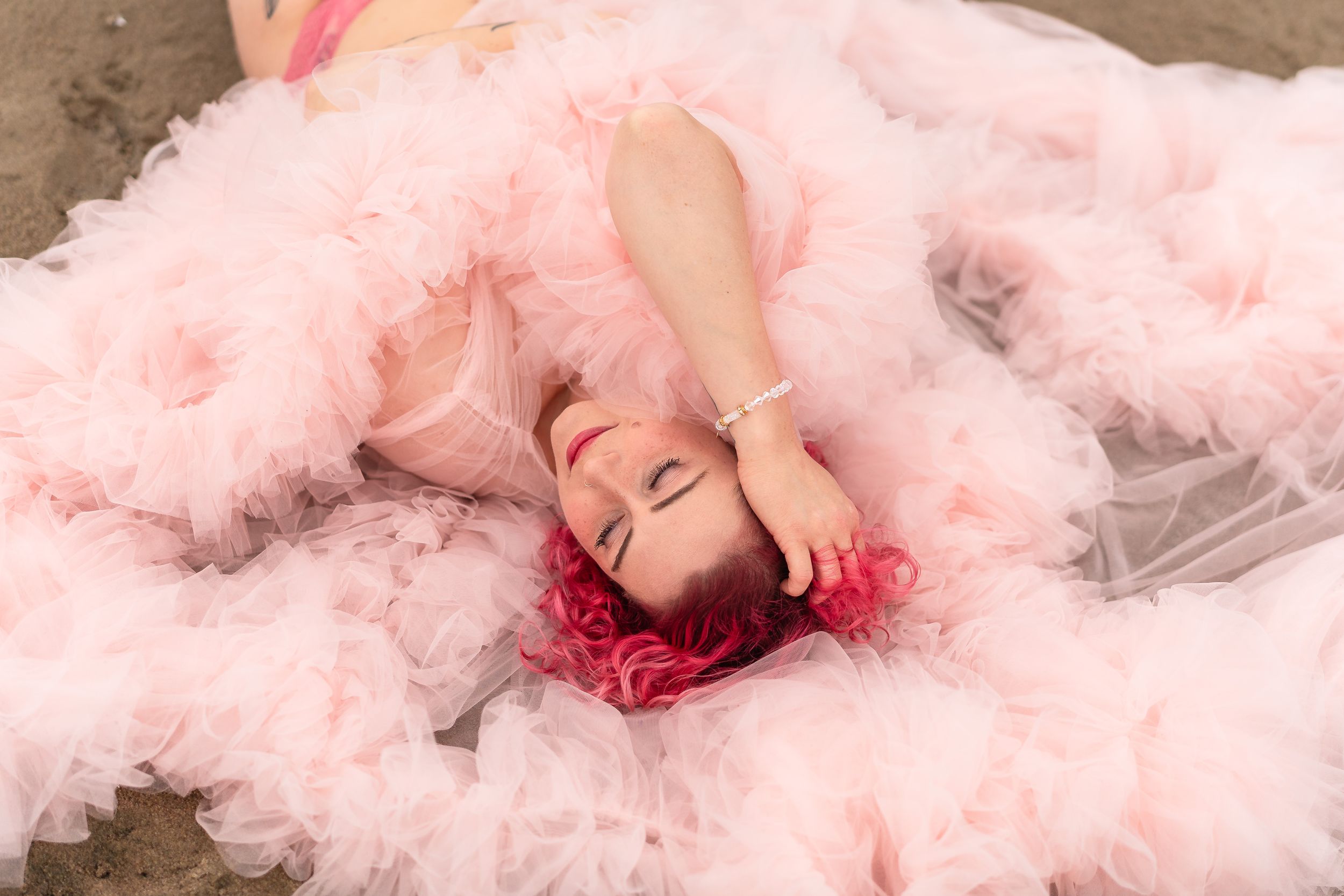 A dreamy portrait of someone lying on pink tulle fabric with vibrant red hair spread out against fluffy material.