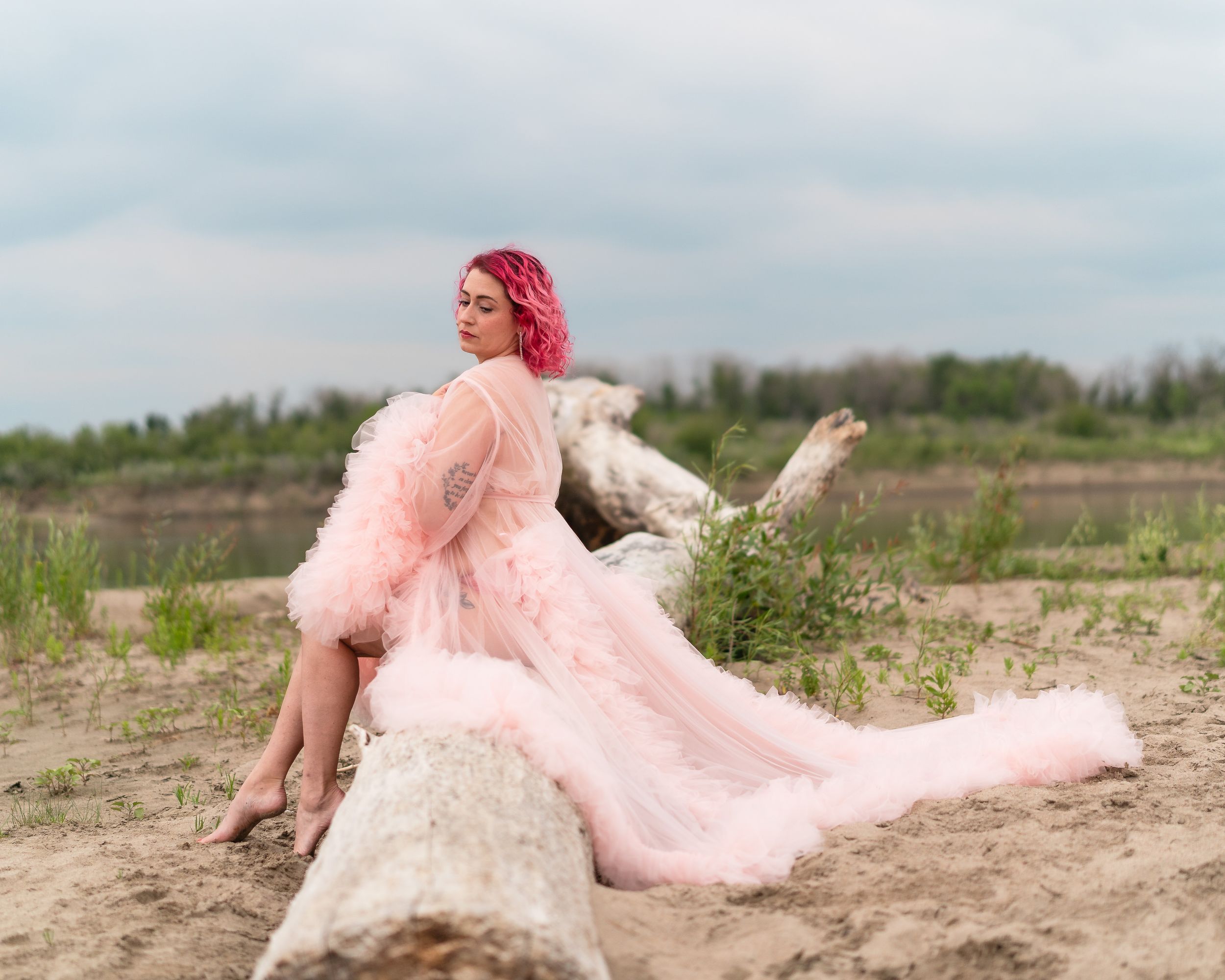 A dreamy portrait shows someone in a pink tulle gown posing gracefully on a fallen log near water.