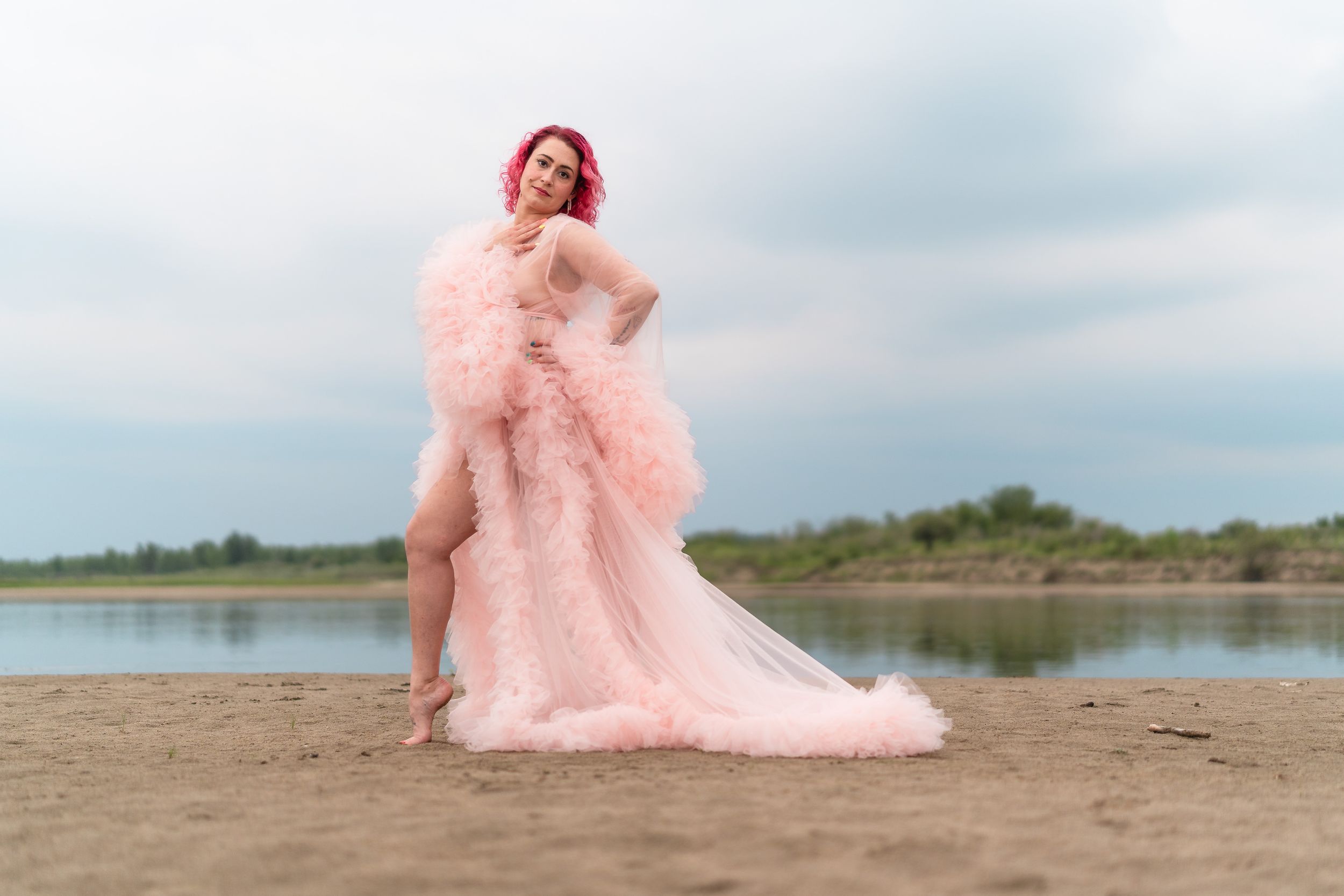 A dreamy fashion shoot on a sandy beach features a flowing pink tulle gown against a cloudy sky.