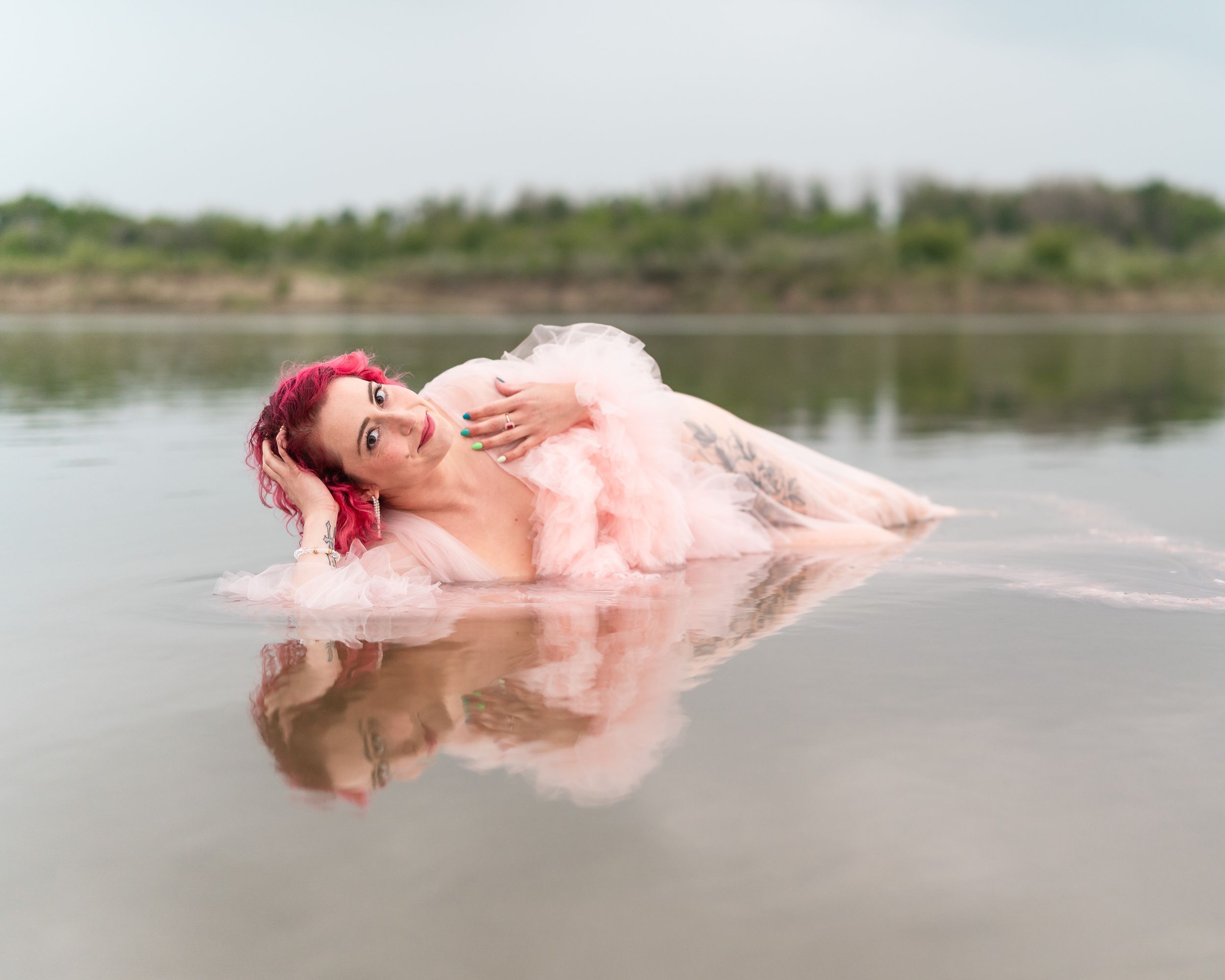 Figure in pink dress floating ethereally in shallow reflective water with pink hair creating a dreamy artistic scene.