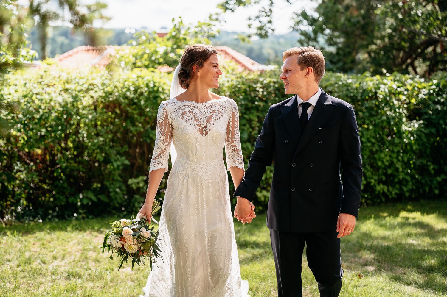 Newlywed couple walks and laughs together in sunny garden while holding a rustic floral bouquet.
