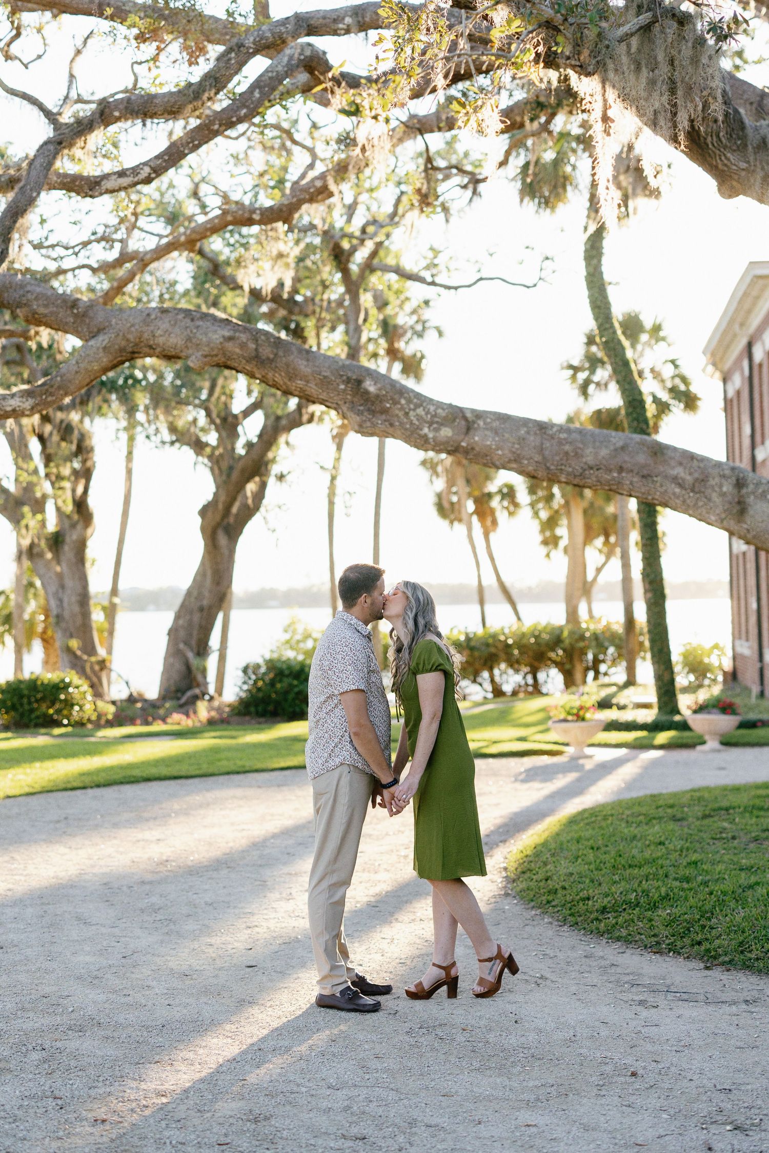 A romantic couple walks and embraces under moss-draped oak trees in a scenic outdoor setting at sunset.