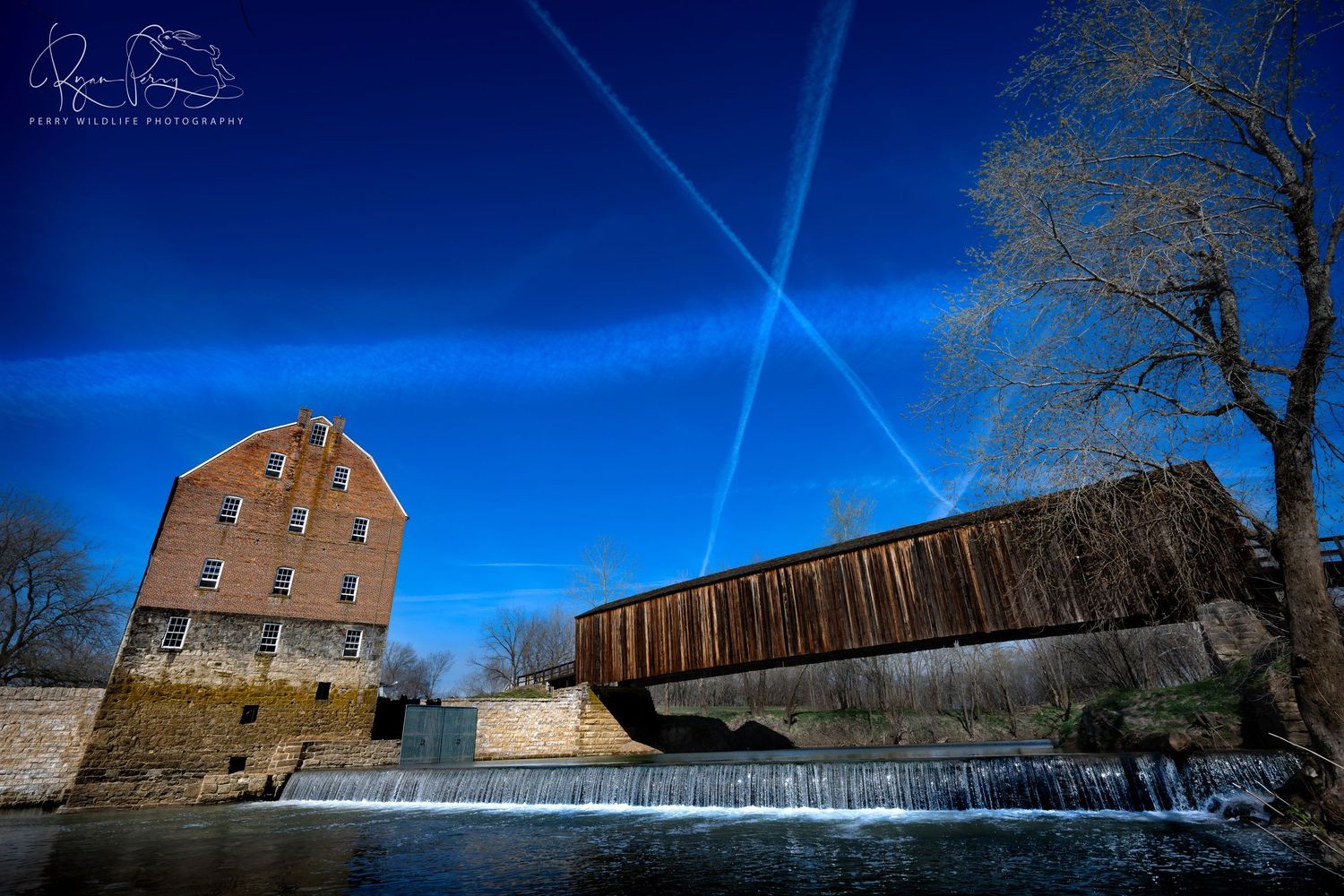 Old brick mill and covered wooden bridge span a cascading waterfall under crossing contrails in a deep blue sky.
