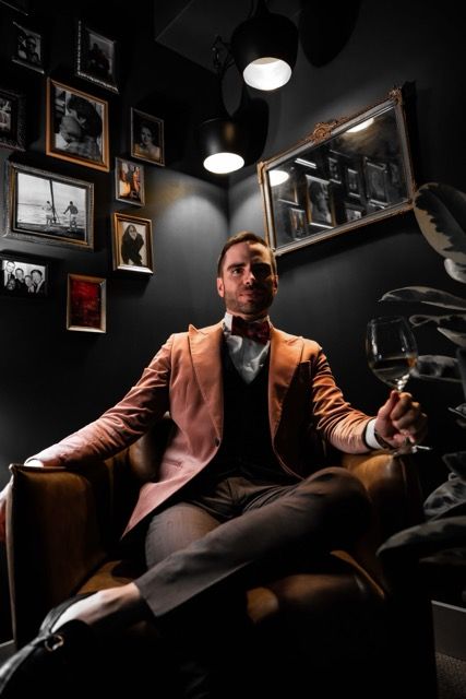 A person in a brown suit jacket poses dramatically in a dark room with hanging pendant lights and framed photos.