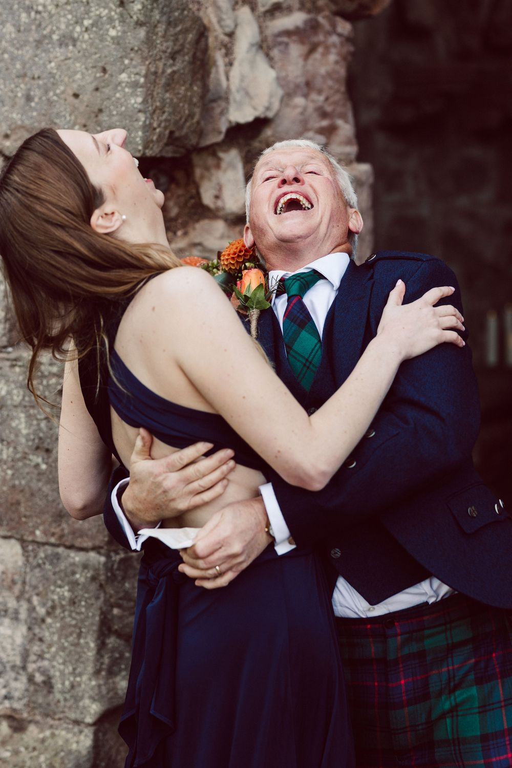 A joyful couple laughs together at a wedding, with the groom wearing a traditional Scottish kilt and navy jacket.