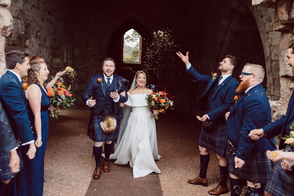 Wedding party guests enjoy a fun candid moment in an outdoor stone archway setting during celebration.