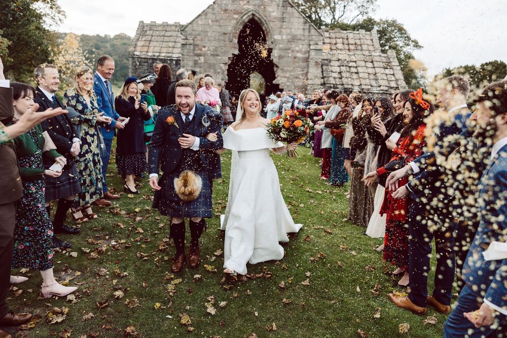 Wedding guests shower newlyweds with flower petals during an outdoor celebration at a rustic stone venue.