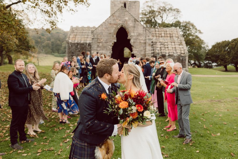 A wedding celebration outside a rustic stone church in autumn with guests gathered on the grass.