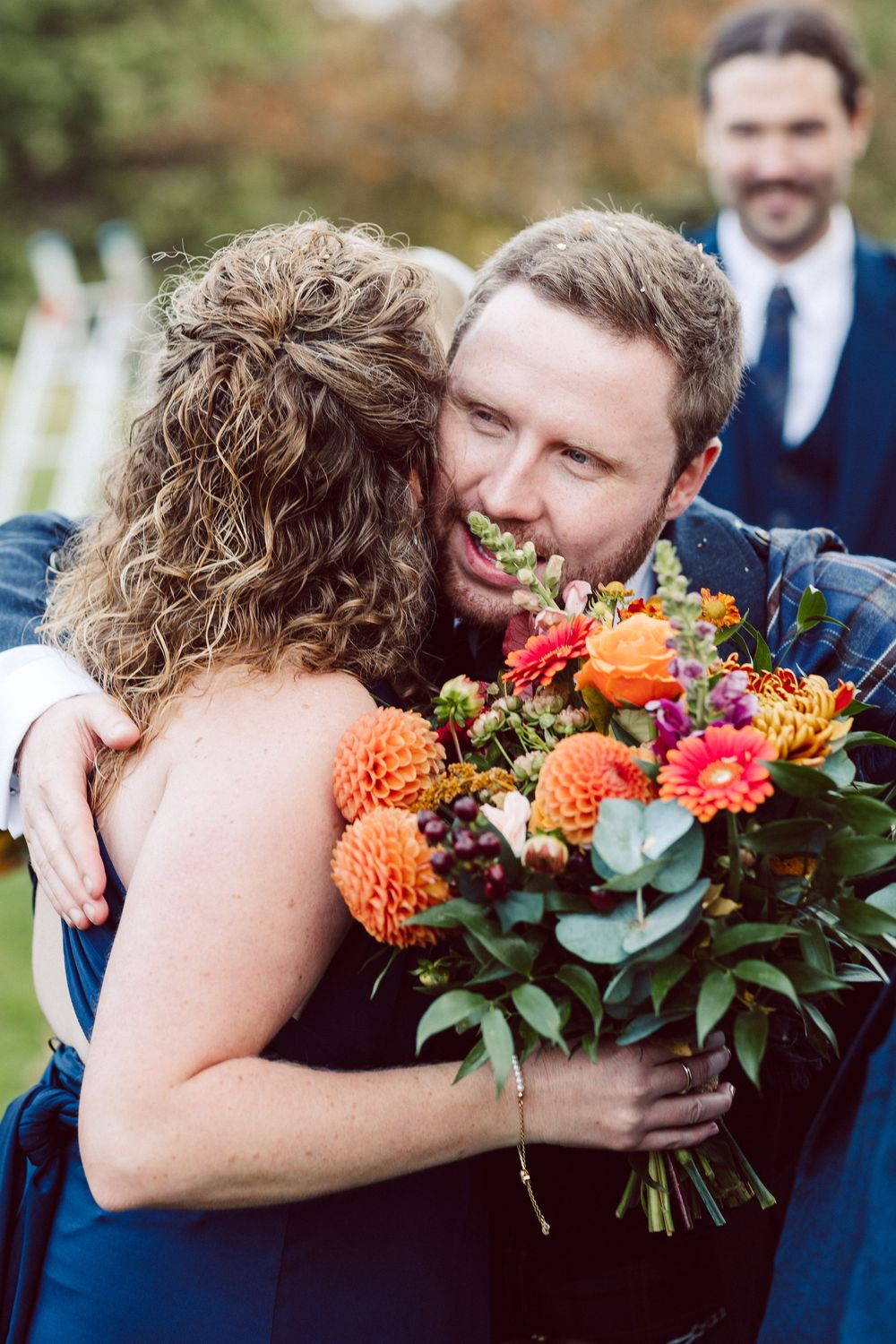 Wedding party members share emotional hugs while holding vibrant orange and red floral bouquets.