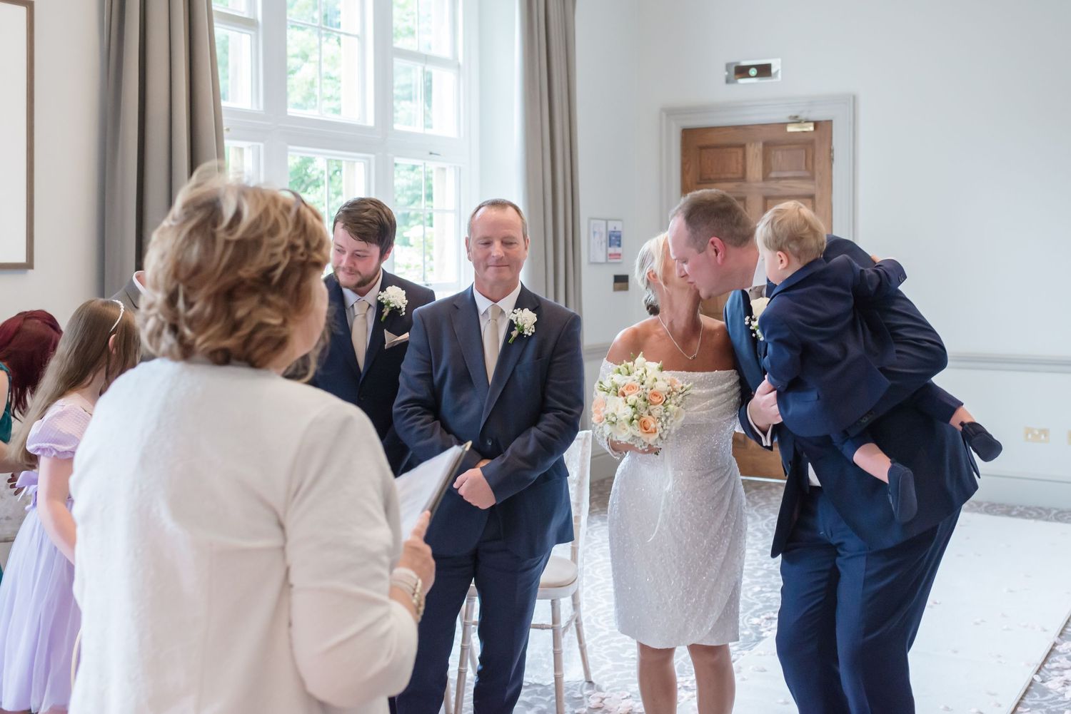 A wedding ceremony in a bright indoor venue with white walls and natural light streaming through windows at DeVere Cranage Estate