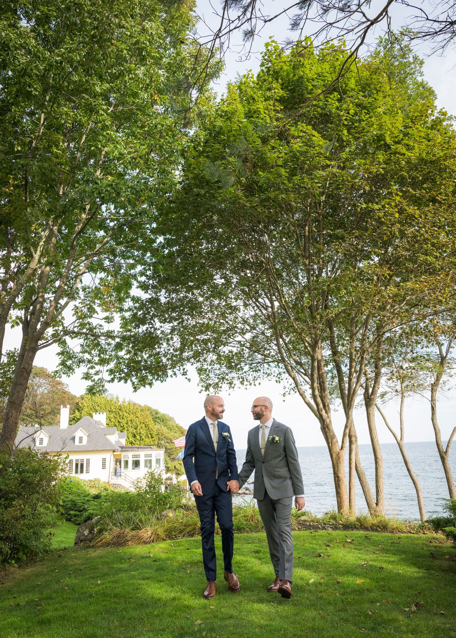 Two people walk and converse on a grassy lawn overlooking a scenic waterfront on a sunny summer day.