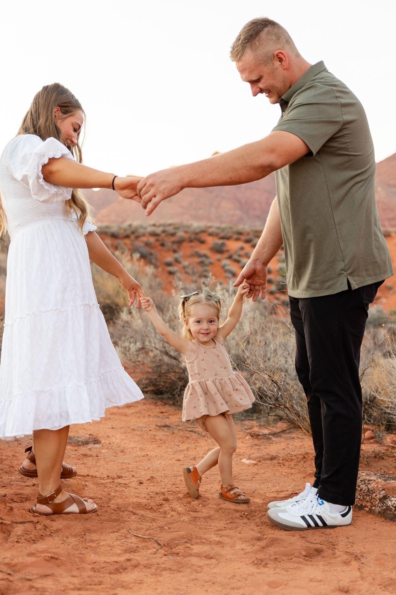 kayenta Ivins in southern utah mom in a long white dress holding hands with dad and a 1.5 year old girl with the red rocks in the background