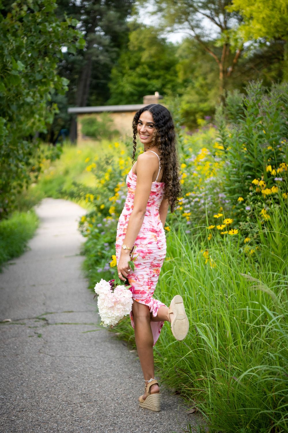 A person in a pink floral dress walks along a winding gravel path bordered by tall wildflowers and lush summer greenery.