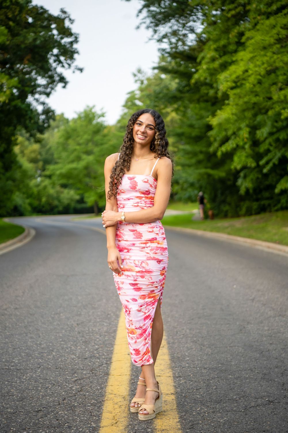 A person in a pink and white patterned dress poses playfully on a curved road surrounded by green trees.
