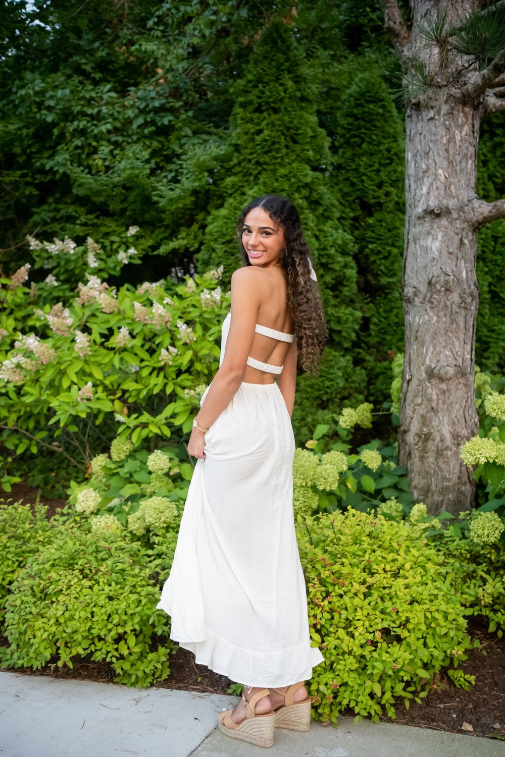 A person in a flowing white sundress poses gracefully in a lush garden setting with tall trees and green foliage.