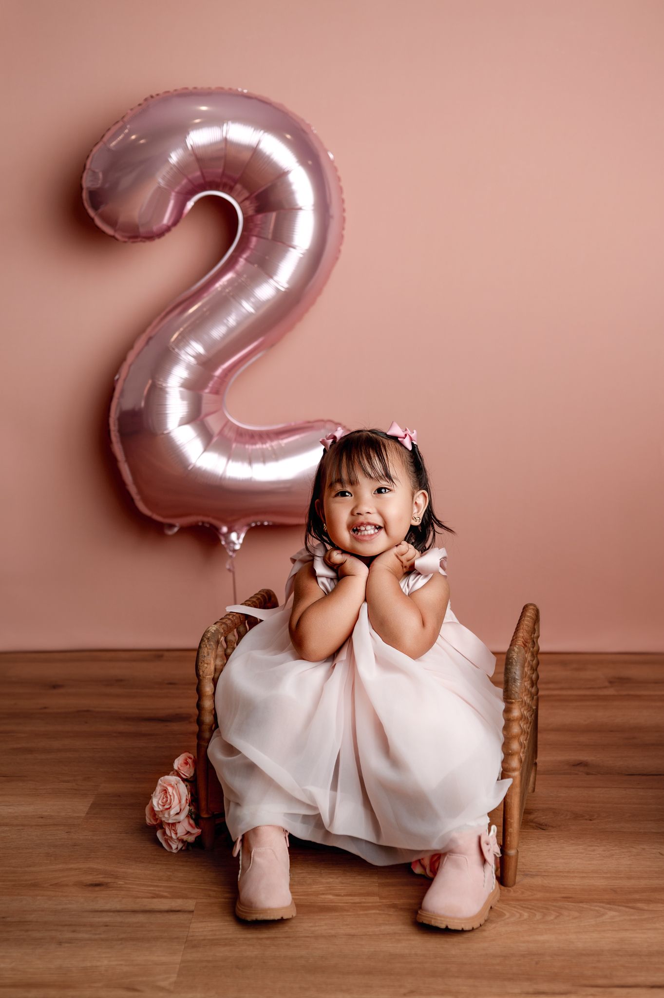 A small child in a white dress sits on a wooden floor with a large rose gold number 2 balloon in the background.