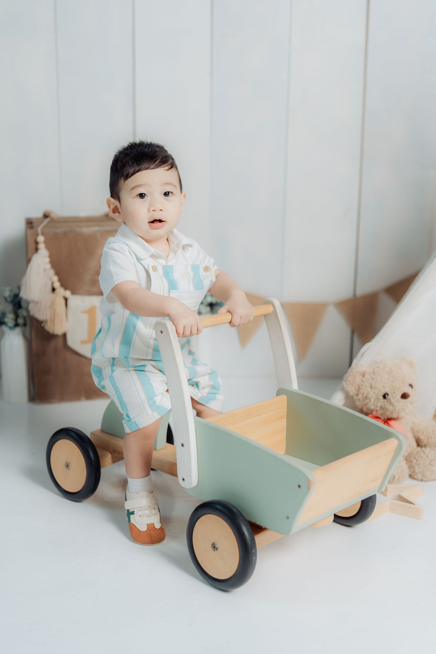 A baby in a mint green outfit plays with a wooden push cart toy while a teddy bear sits nearby.
