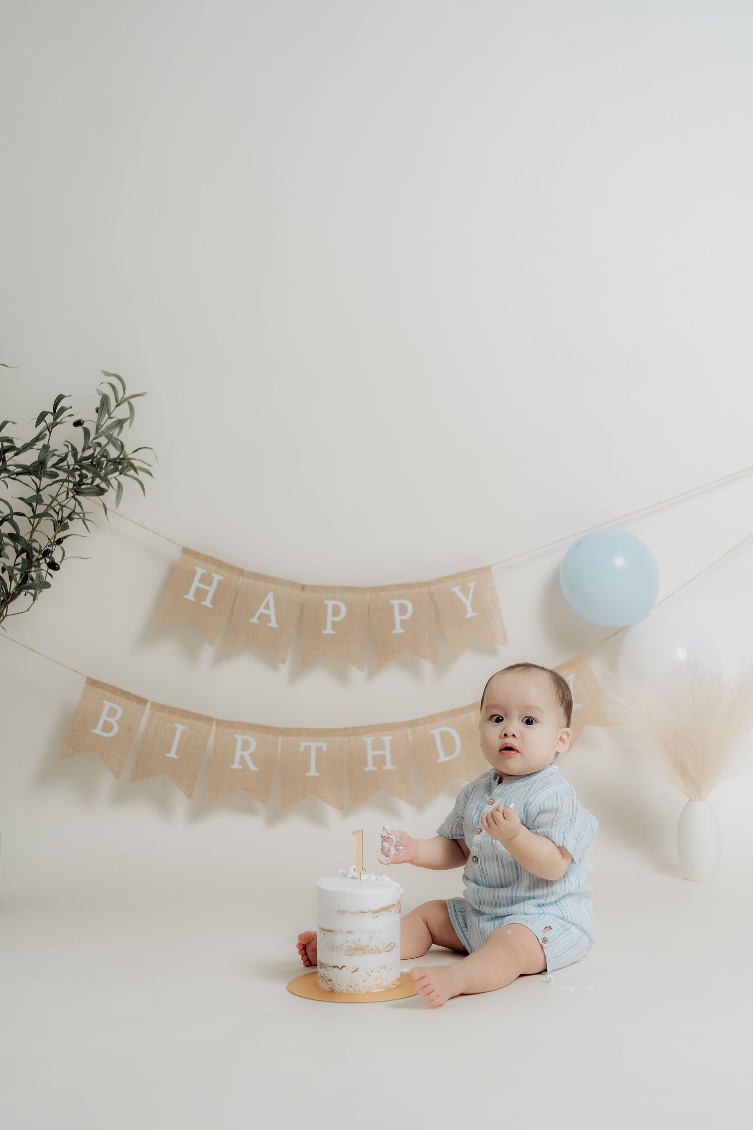 Baby in blue outfit lying on floor with birthday banner and cake for cake smash photoshoot.