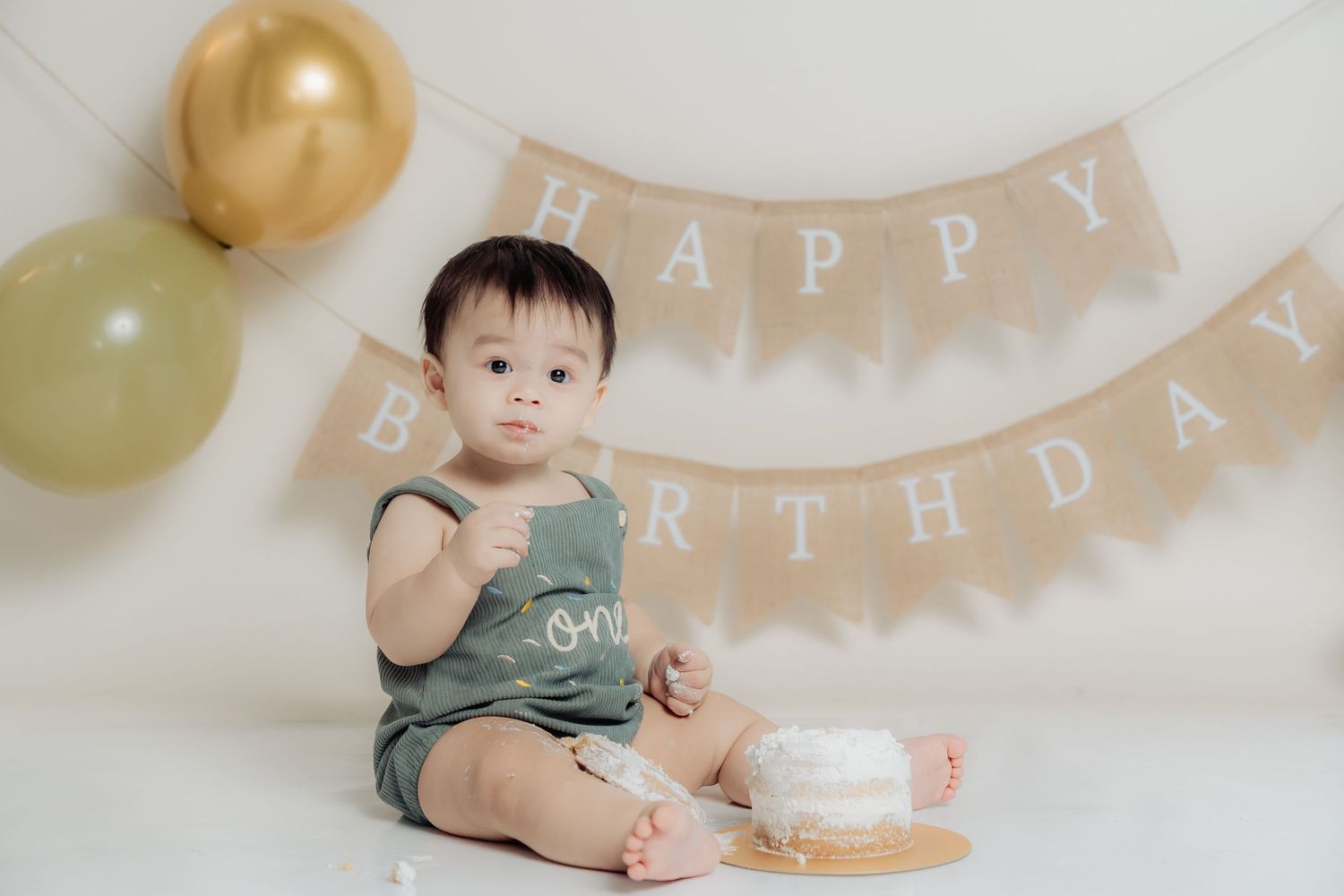 Minimalist first birthday setup with gold balloons and happy birthday banner against white backdrop.