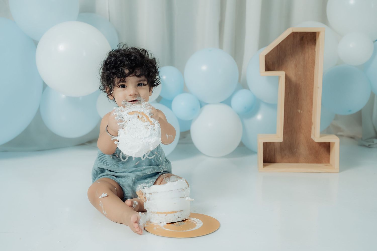 A baby in a blue and white balloon-filled room smashes a cake during a first birthday cake smash photo session.