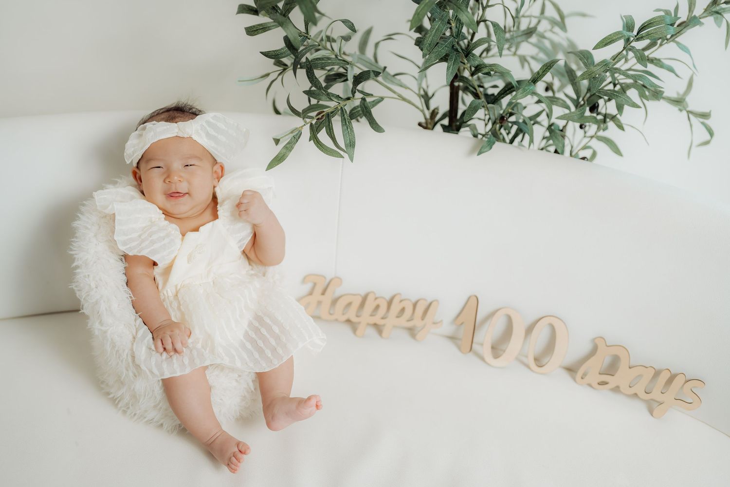 A baby in white clothing lies on a white backdrop with decorative text reading 'Happy 100 Days' and green foliage above.