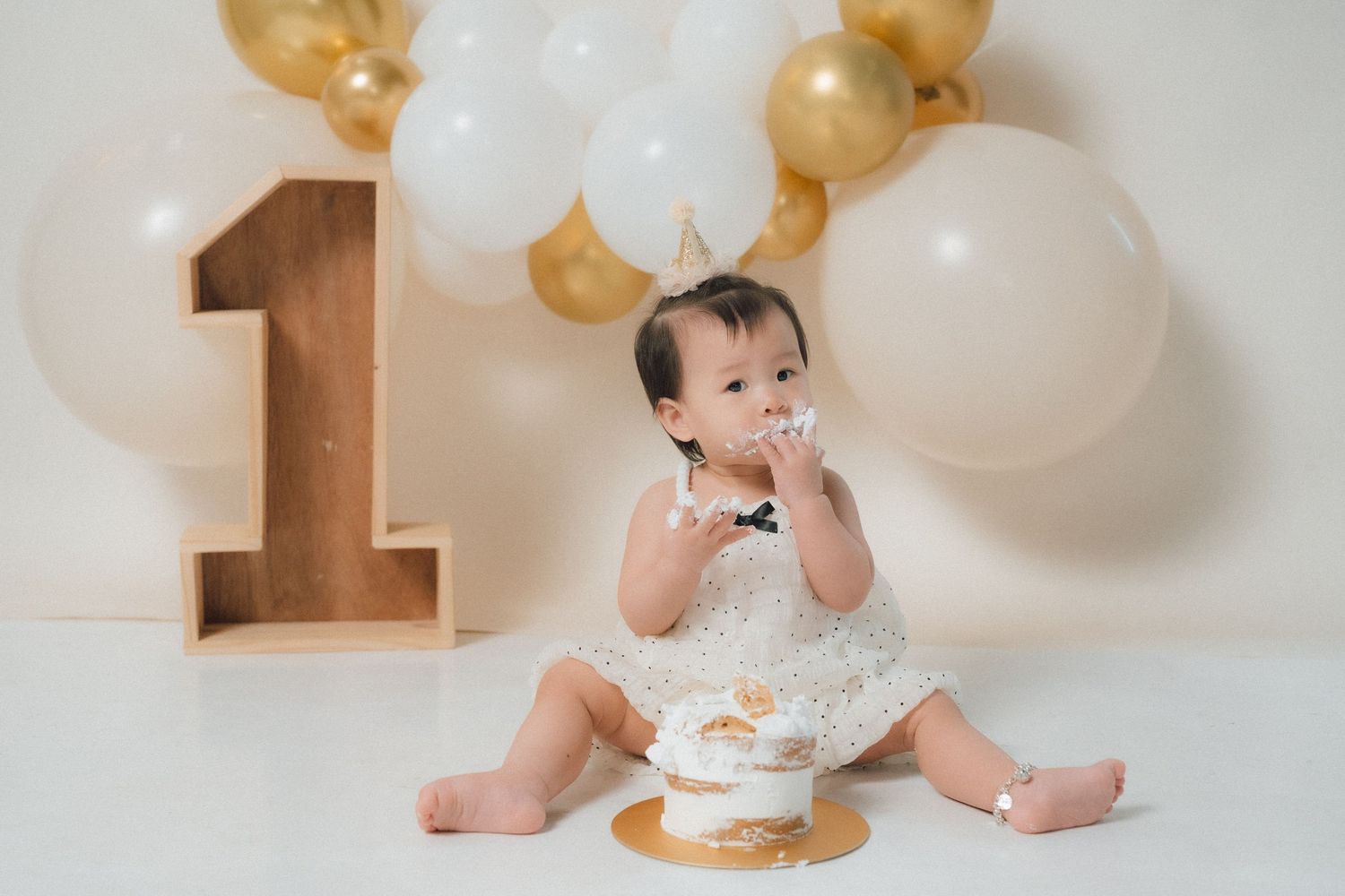 Baby in lace dress having cake smash photoshoot with gold and white balloon decorations.