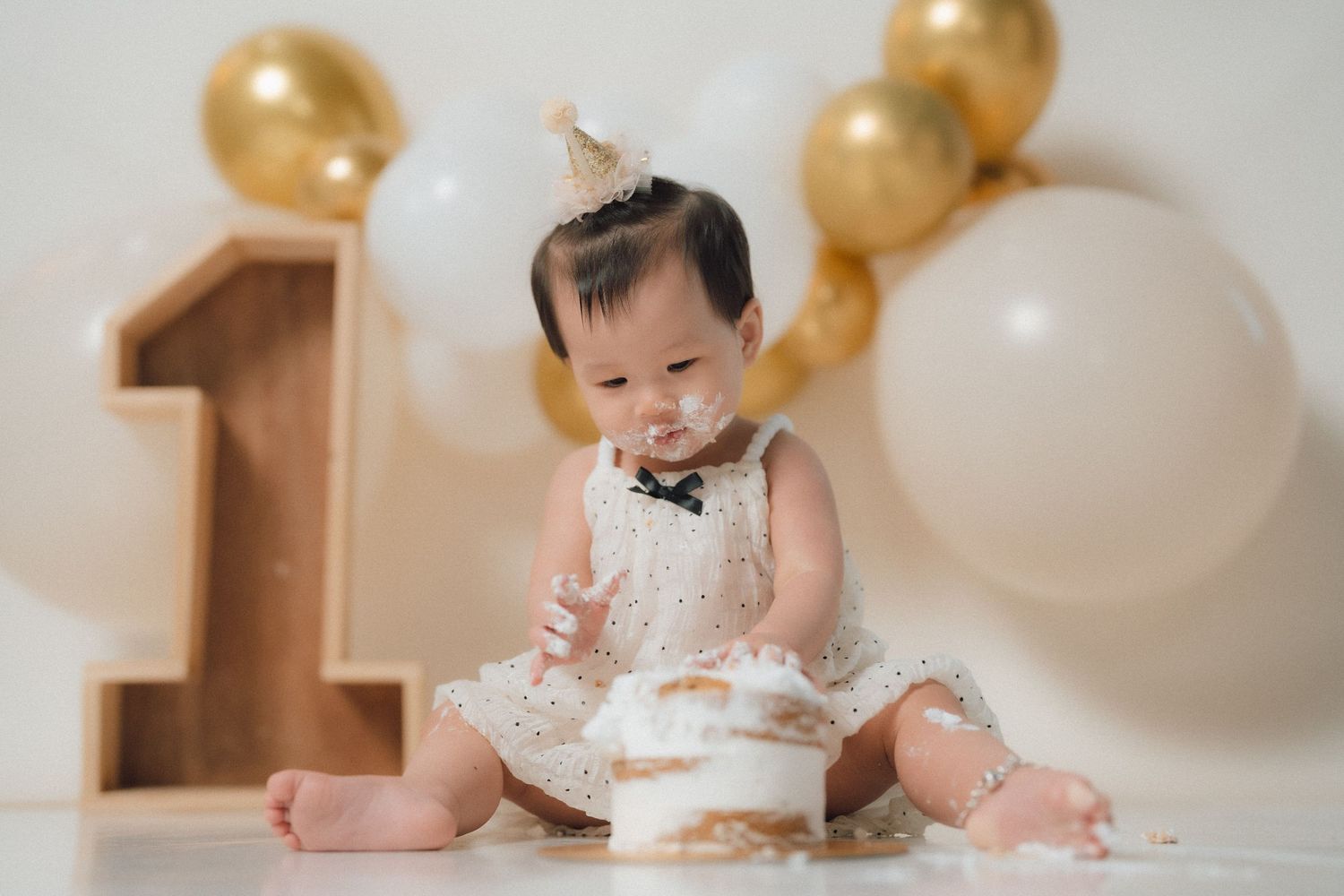 First birthday cake smash session with baby in white dress and gold number one decoration.