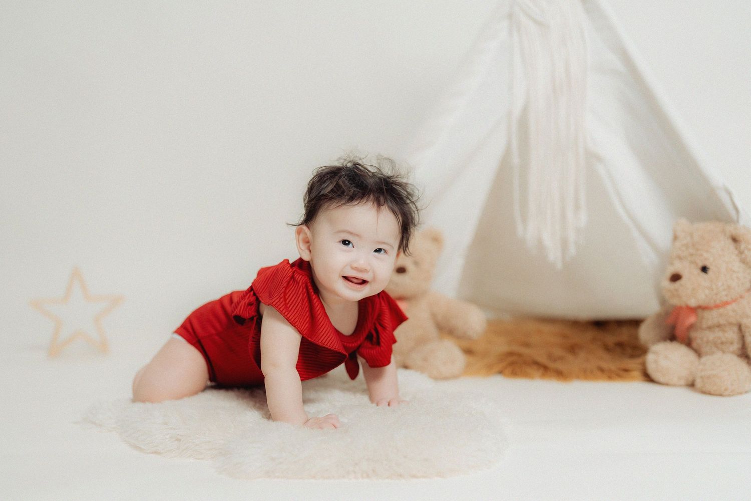A small figure in a red outfit crawls on a white blanket next to a teepee tent and plush teddy bear.