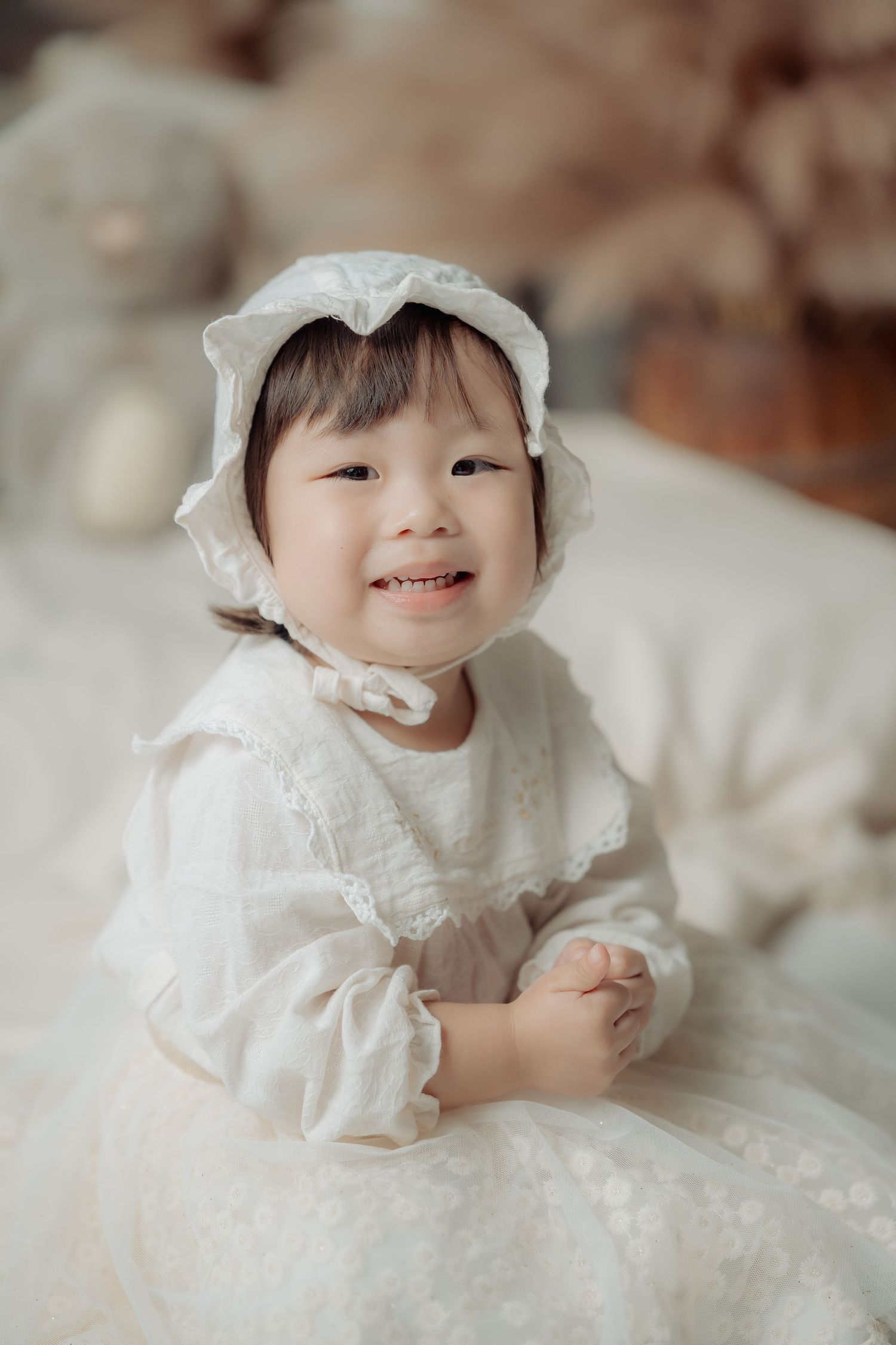 Baby wearing white lace dress poses on cream colored background.