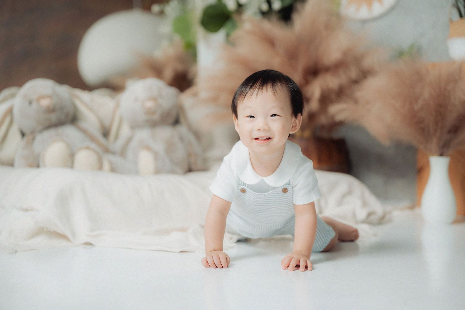 A small child in light blue overalls crawls on a white floor with plush toys and decorative elements in the background.