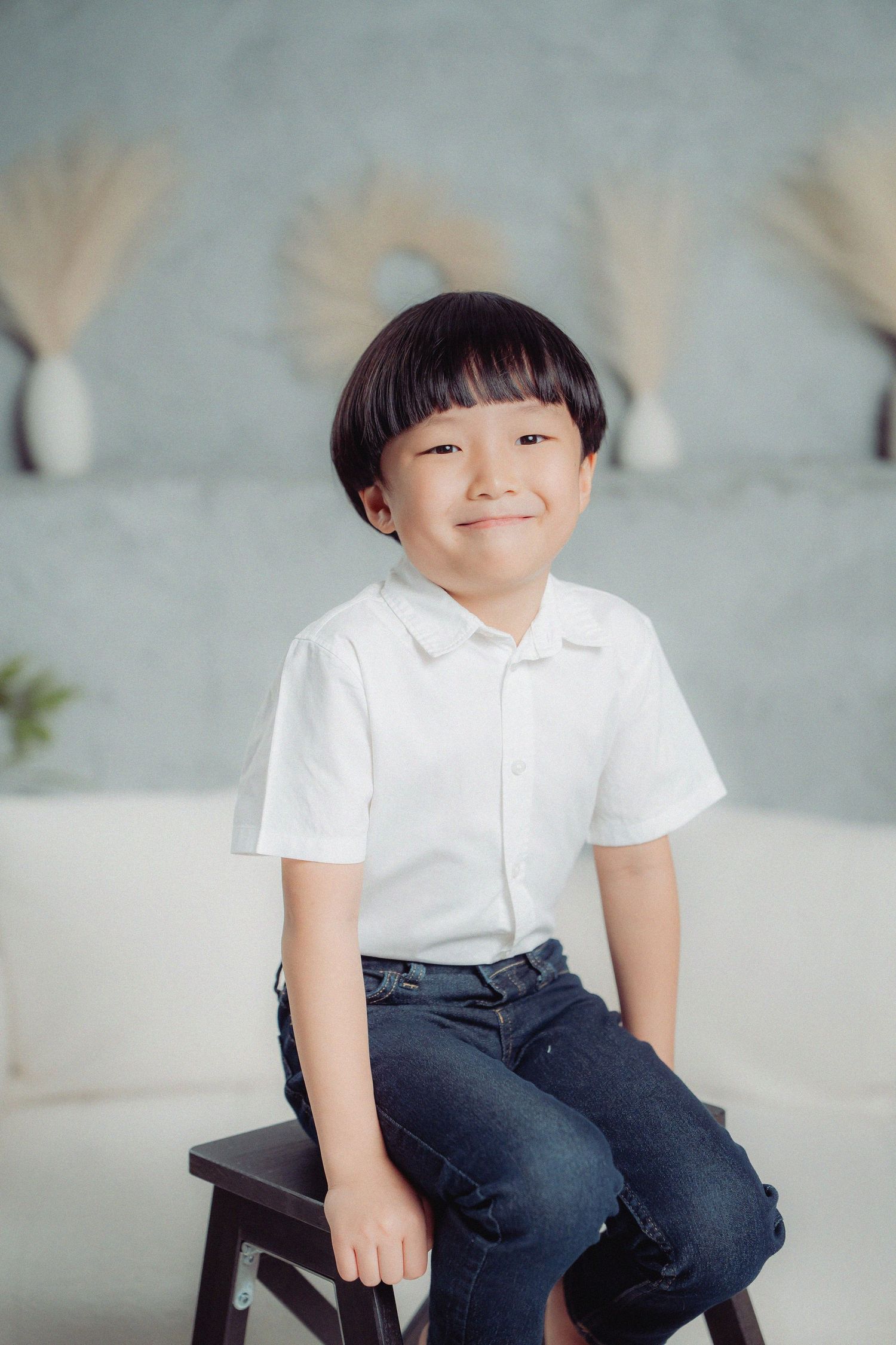 Child in white polo shirt and jeans sits on dark wooden stool against light gray wall with pampas grass decor.