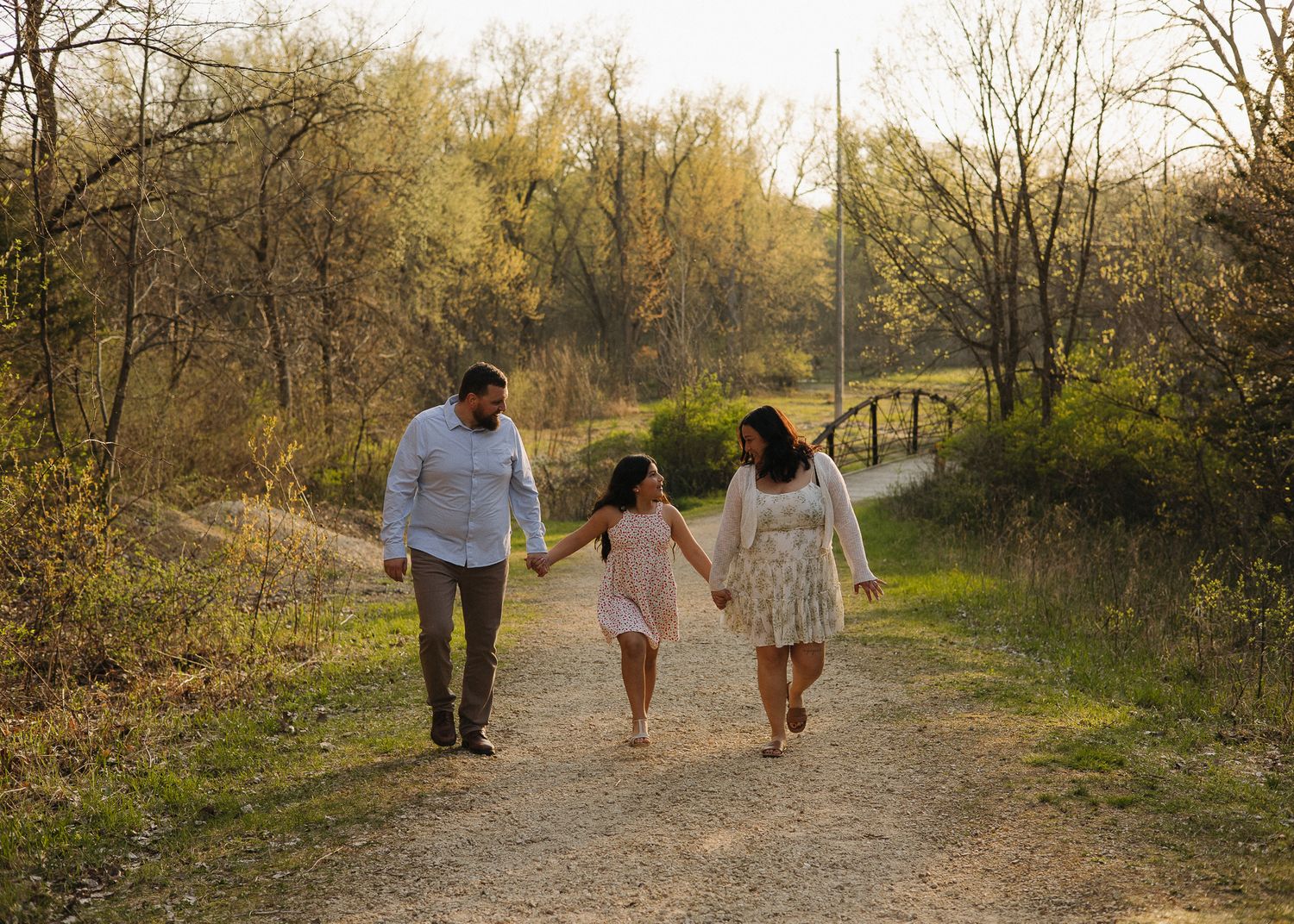 Happy family of three holding hands and walking along a tree-lined path in golden light.