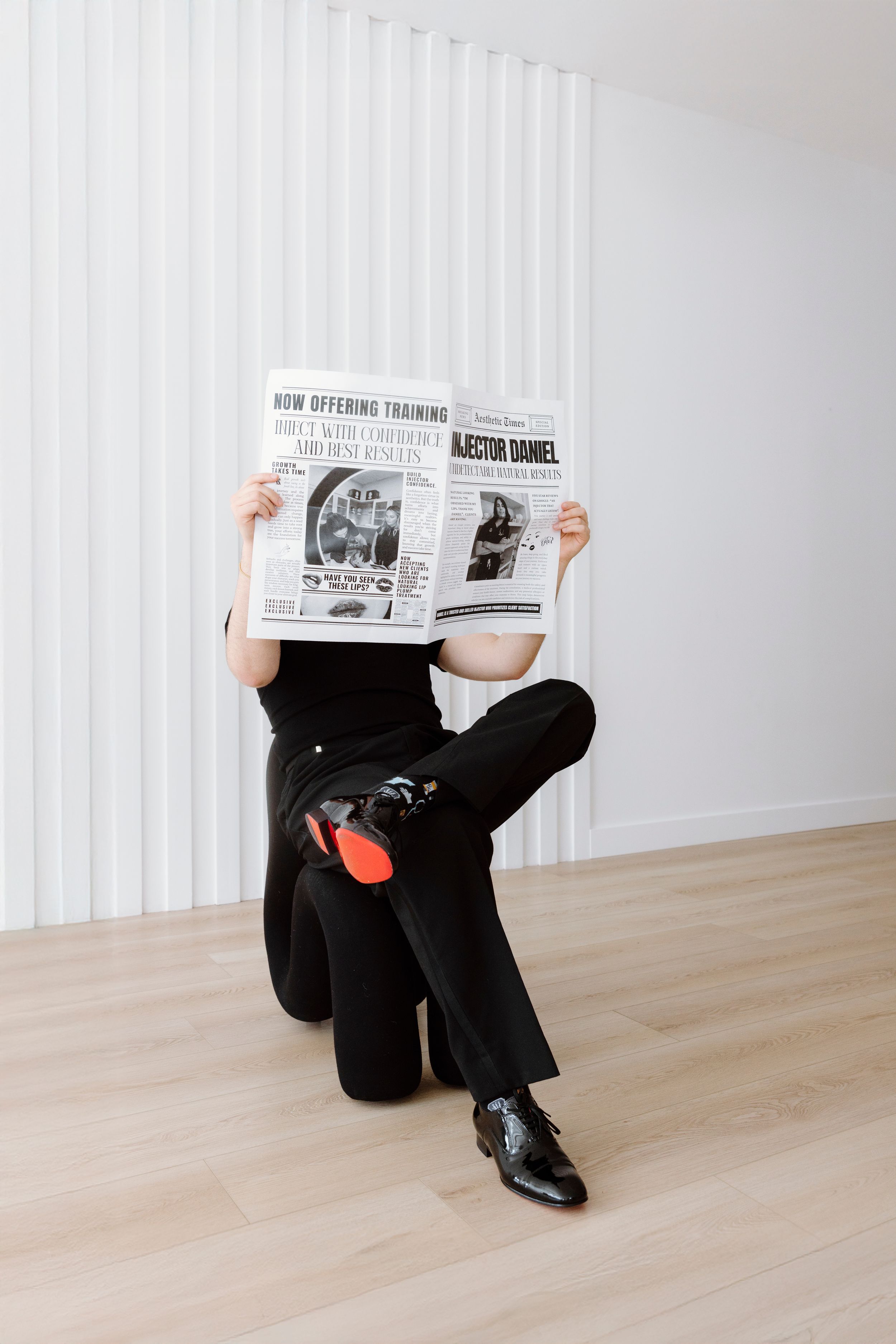 Person in black attire sitting on wooden floor while reading newspaper against white wall.